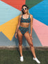 Two-piece high-waisted bikini set featuring an all-over print of blue skulls with purple bows and multicolored stars on a dark background. Includes a deep V-neck bikini top with wide straps and matching high-rise bottoms. Made from soft, stretchy fabric with a smooth black inner lining. The design combines bold, edgy graphics with a flattering retro-inspired silhouette.